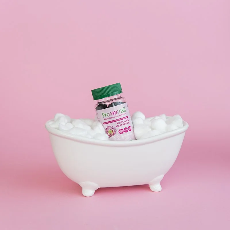 A small bottle labeled 'Pro menstru' resting inside a white bathtub filled with white foam, against a pink background.