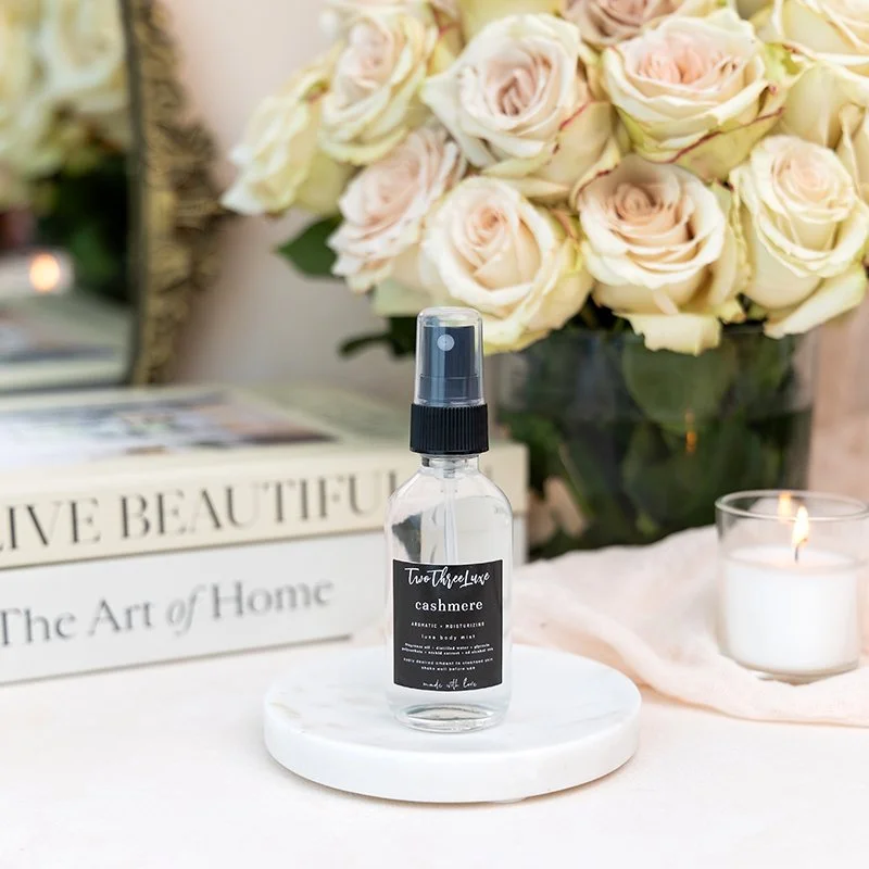 A clear spray bottle labeled 'Two Three Luxe cashmere.' It is placed on a white circular platform in front of a bouquet of light pink and cream roses, a candle, and a decorative arrangement on a table.
