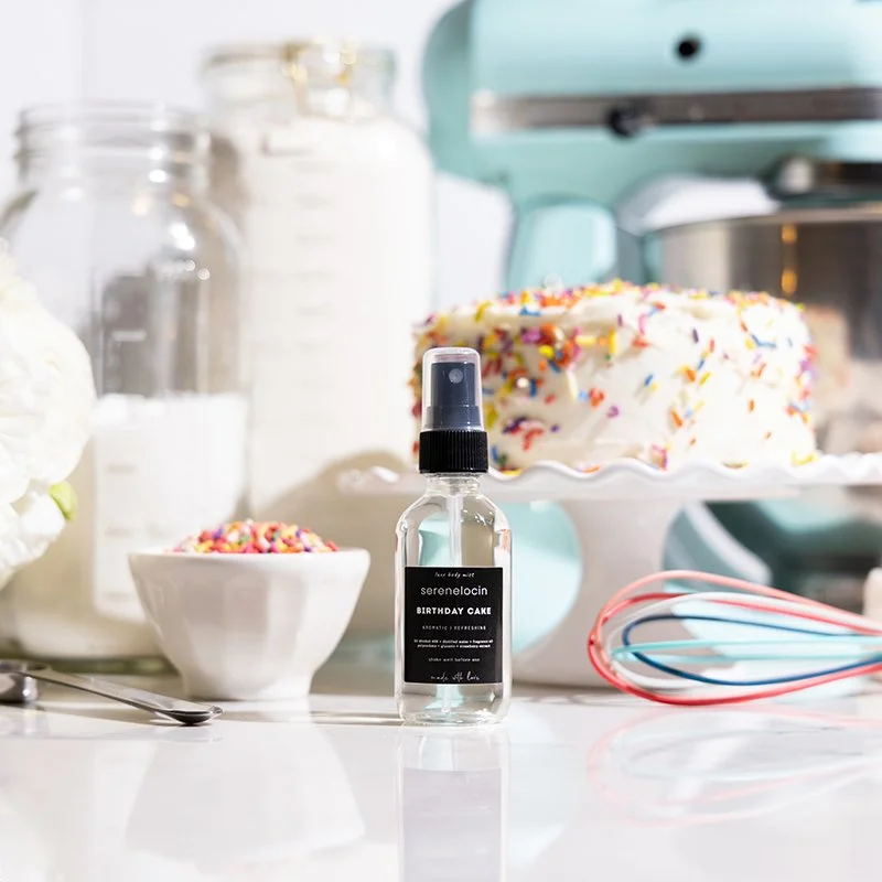 A spray bottle labeled 'Birthday Cake' standing on a white countertop with birthday cake, sprinkles, mixing bowls, a stand mixer, and baking ingredients in the background.