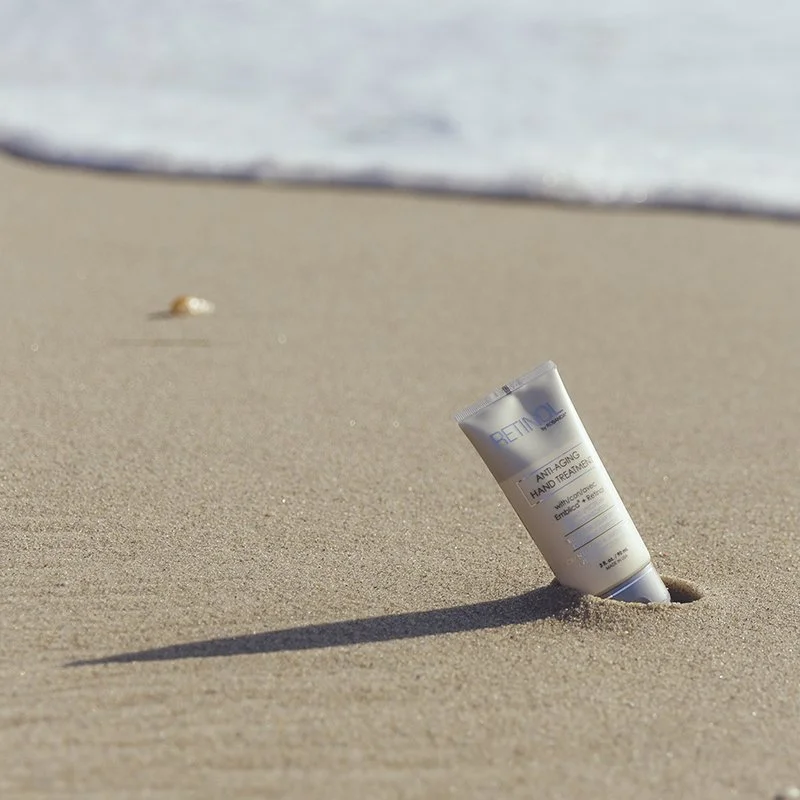 A tube of Retinol anti-aging hand treatment partially buried in sandy beach with a shell in the background and the ocean in the distance.