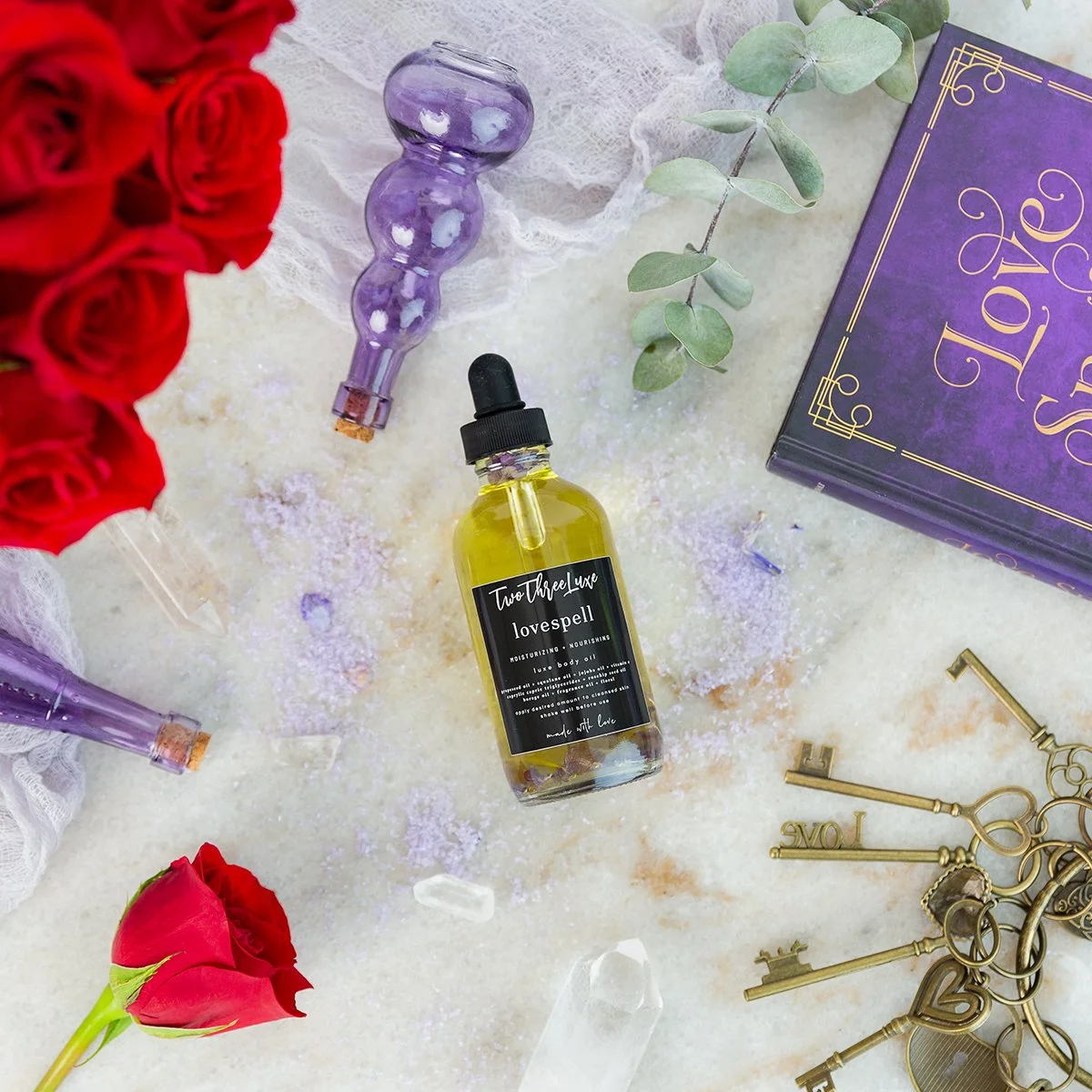 A flat lay of a yellow bottle labeled 'lovespell' from Two Three Luxe, surrounded by red roses, purple glass objects, a eucalyptus branch, a purple love-themed box, clear crystals, and gold keys on a light surface.