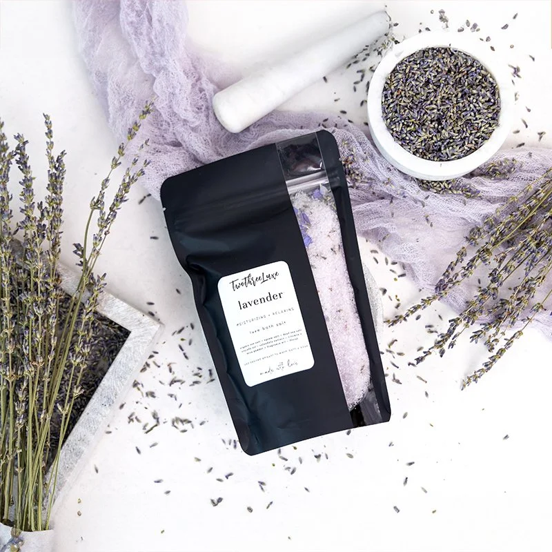 Lavender-scented bath salts in a black resealable bag, surrounded by dried lavender sprigs, a mortar and pestle, and a bowl of dried lavender flowers, on a white surface with lavender fabric.