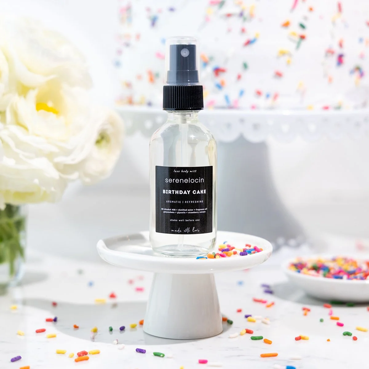 A clear spray bottle labeled "serenelocin BIRTHDAY CAKE" with a black label, placed on a small white pedestal, surrounded by colorful confetti and white flowers, in a bright setting.