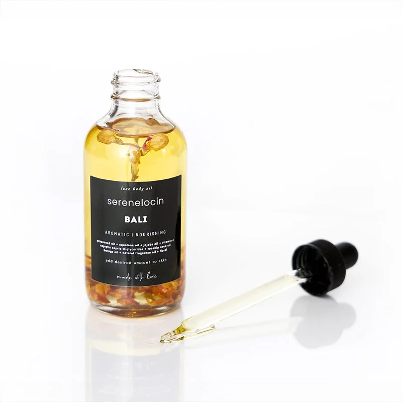 A bottle of balancing body oil labeled 'Serenelocin Bali Aromatic Nourishing' with flower petals inside, and a dropper lying next to it on a white surface.
