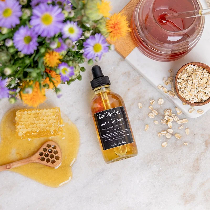 A flat lay of natural skincare or bath products, including a dropper bottle labeled 'oat + honey,' a honeycomb with honey on a wooden honey dipper, a jar of honey, and a bowl of oats, alongside a bouquet of purple and yellow flowers on a white surfac