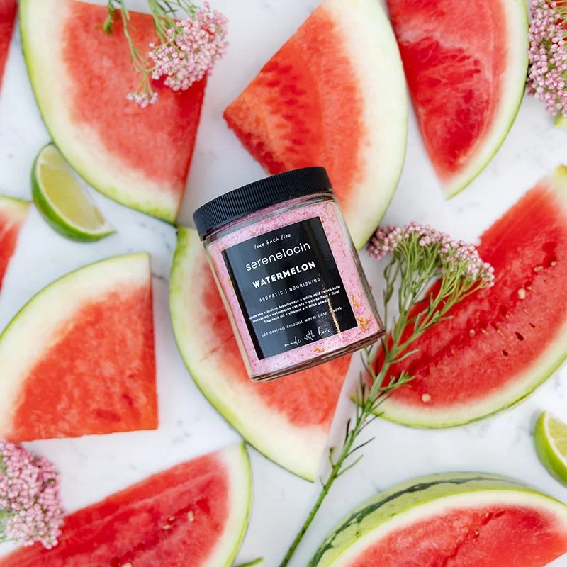 A jar of sereno.locin watermelon face mask surrounded by sliced watermelon pieces, lime wedges, and small pink flowers on a white surface.