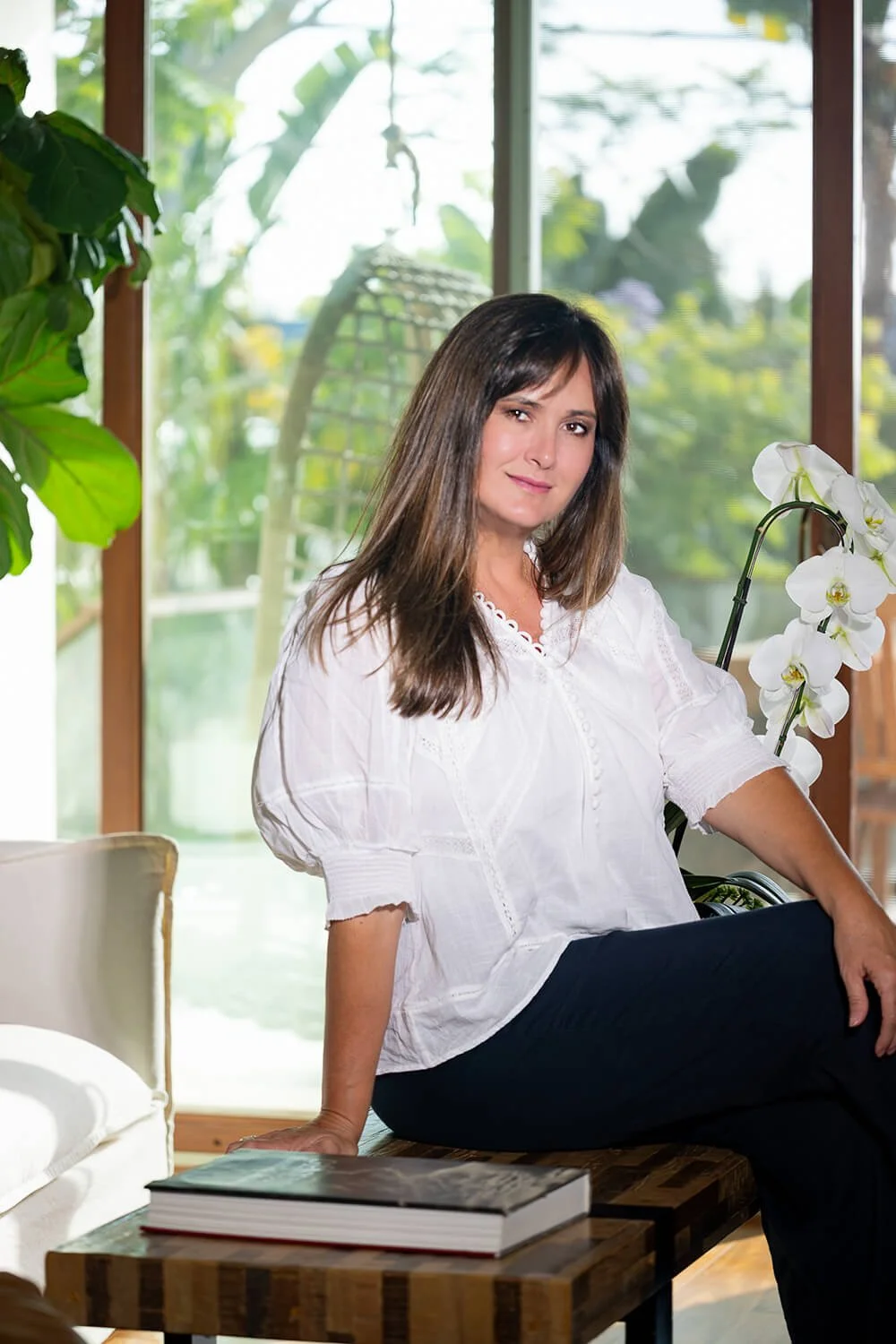 A woman with long brown hair, wearing a white blouse and black pants, is sitting on a wooden bench indoors near large glass windows with greenery outside. There is a white orchid plant beside her and a book on the table in front of her.