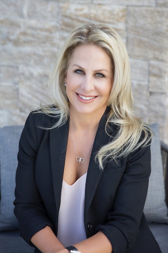 A smiling blonde woman with blue eyes wearing a black blazer and white top sitting on a couch outdoors.