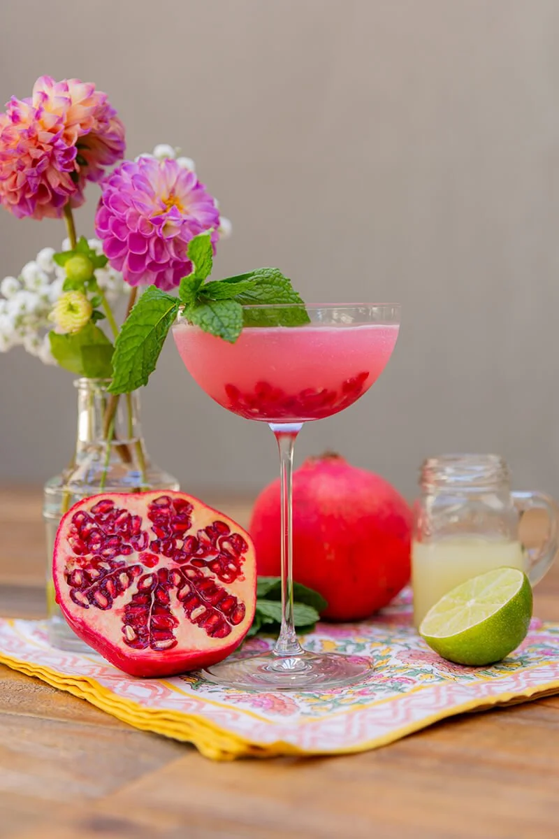 A pink pomegranate cocktail garnished with fresh mint leaves, with a halved pomegranate, whole pomegranate, lime, and a jar of lemonade on a wooden table beside a vase of pink and white flowers.