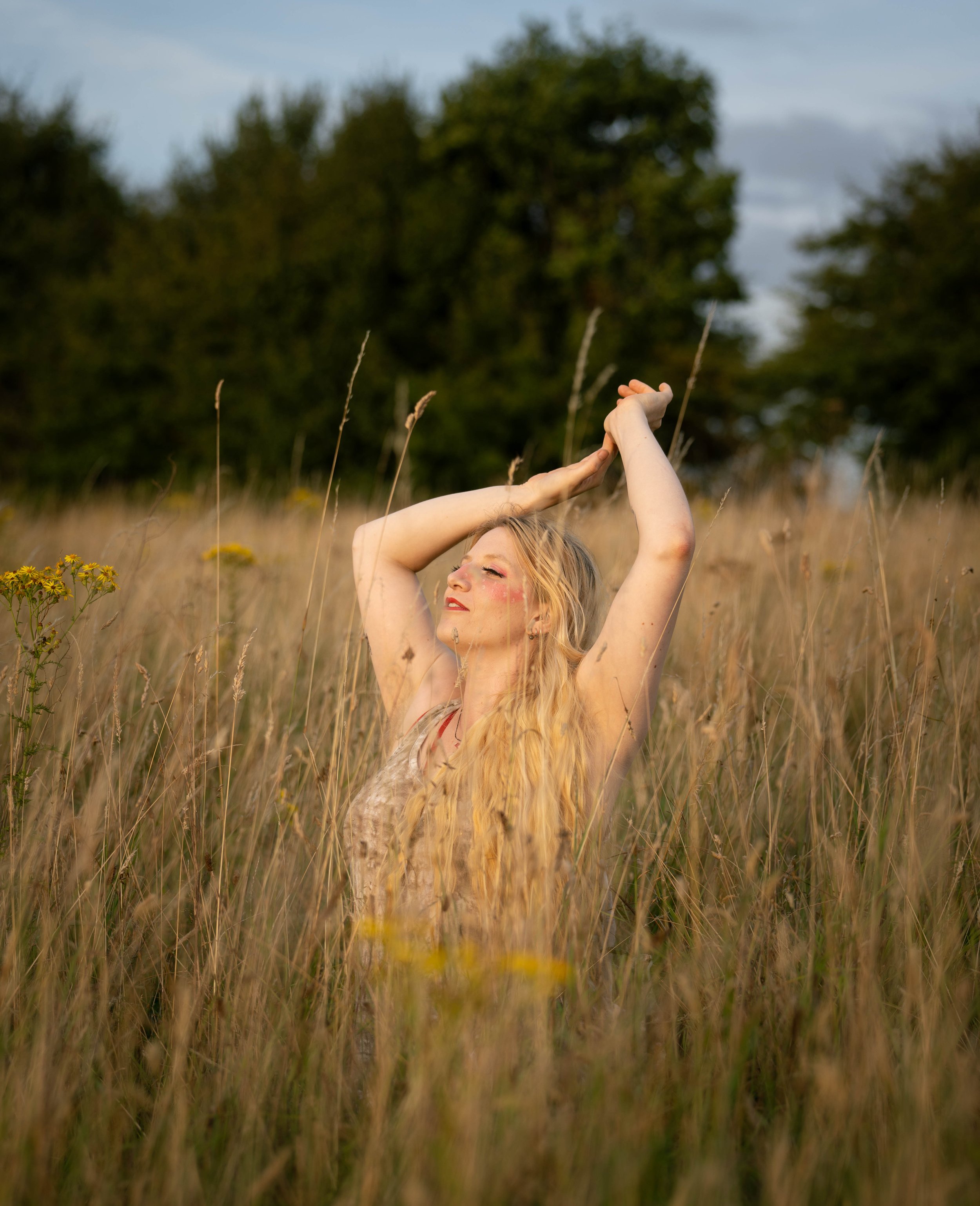 A woman with long blonde hair stands in a field of tall grass, smiling with her eyes closed, arms raised above her head, enjoying the sunlight.