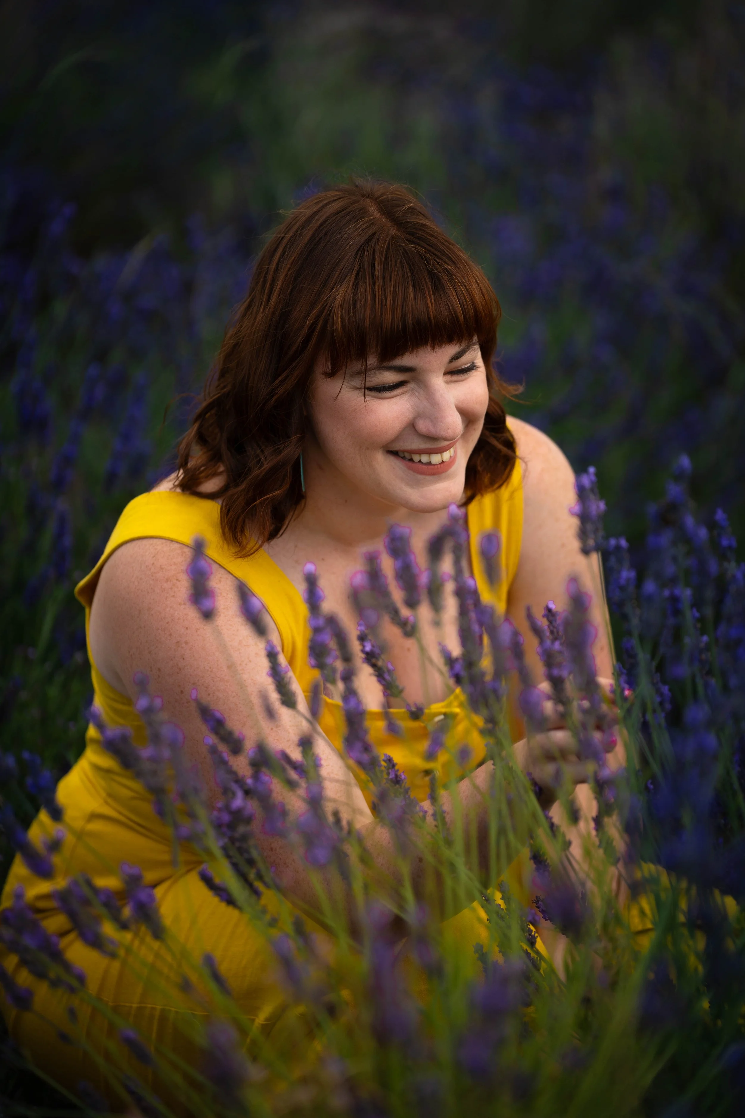 A woman with red hair smiling and sitting in a field of purple lavender flowers, wearing a yellow dress.