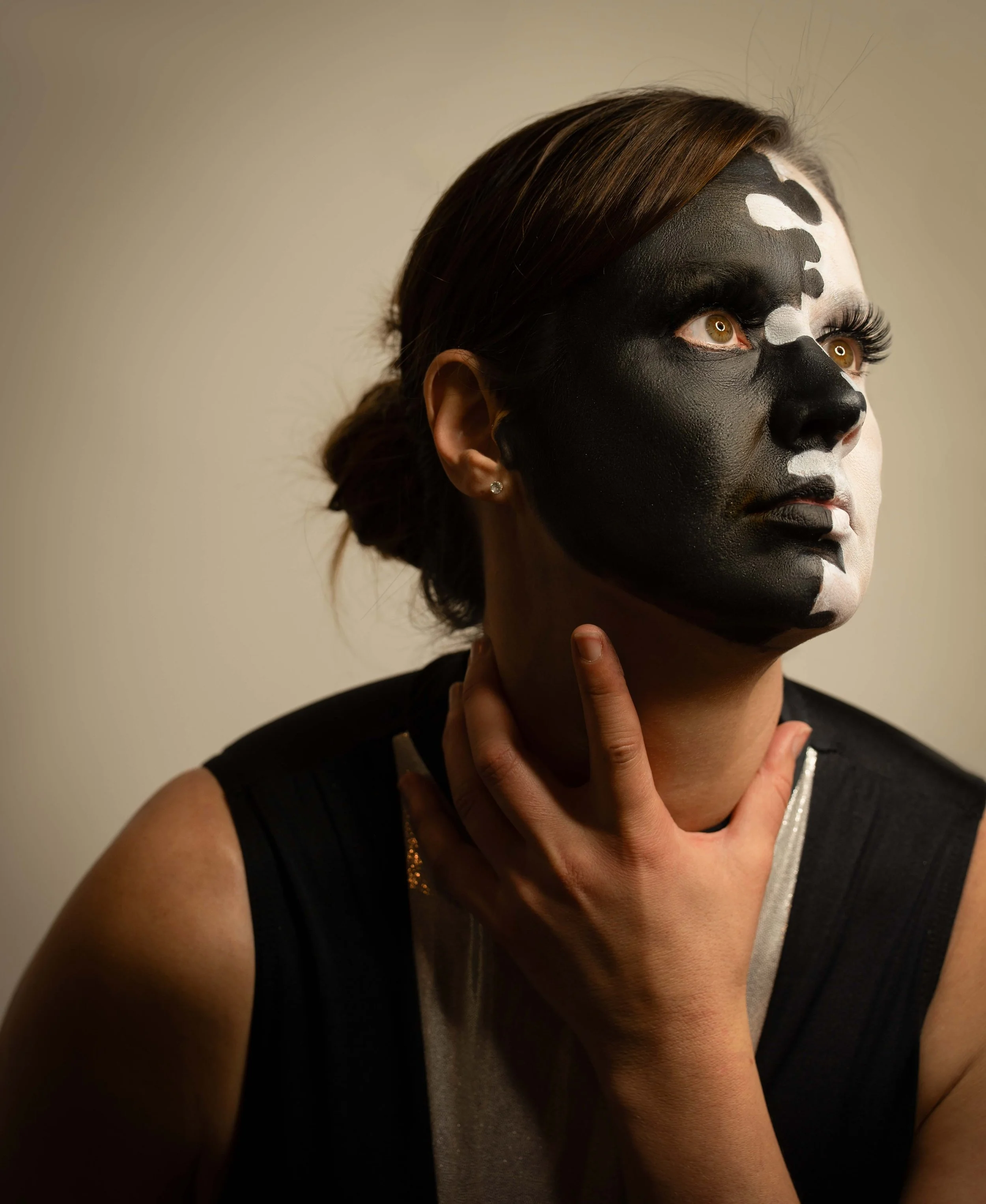 A woman with her face painted in black and white resembling a skull, holding her neck with her hand and looking upward.