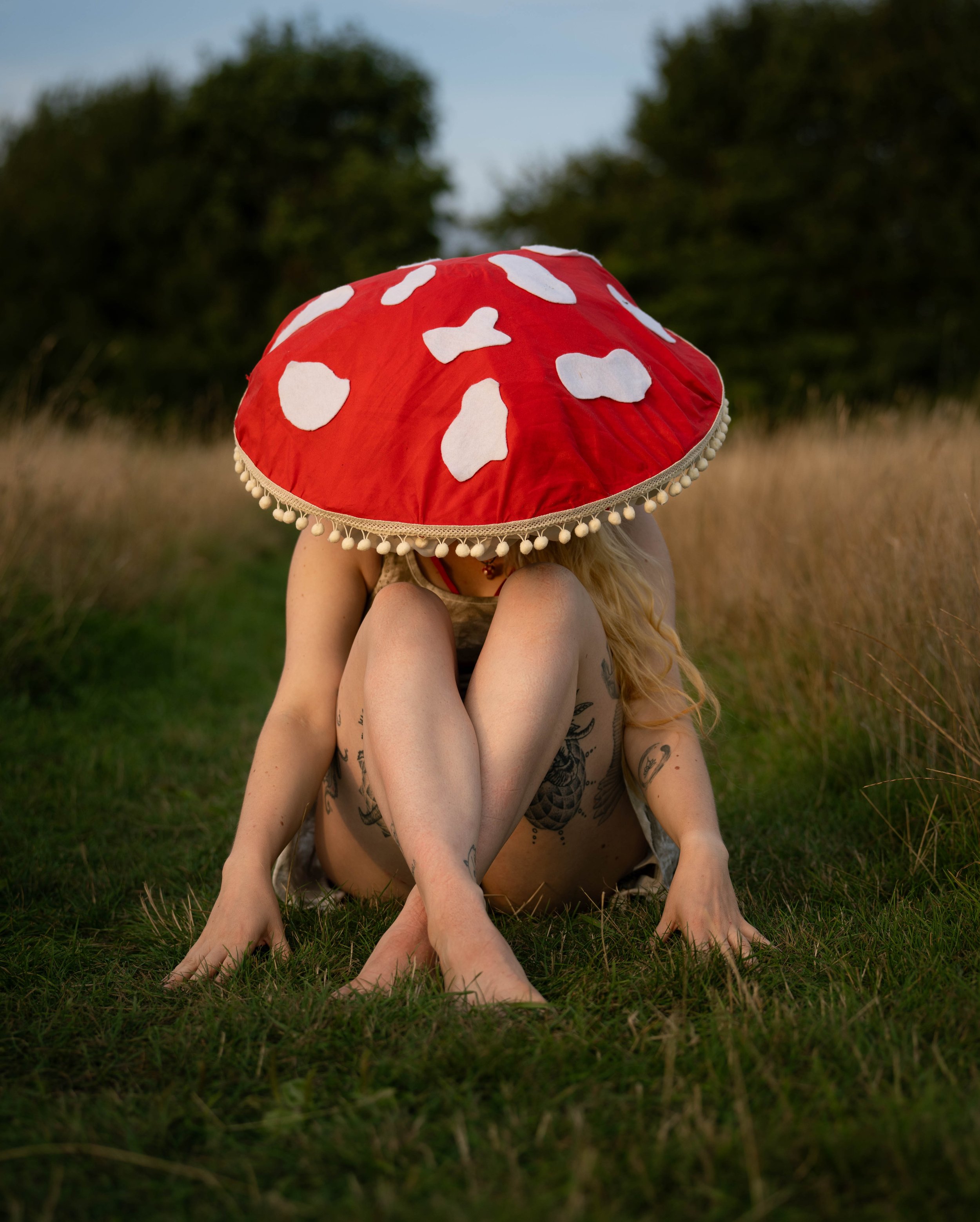 Person sitting cross-legged on grass, wearing a large mushroom-shaped hat with red and white spots, outdoors in a field with trees in the background.