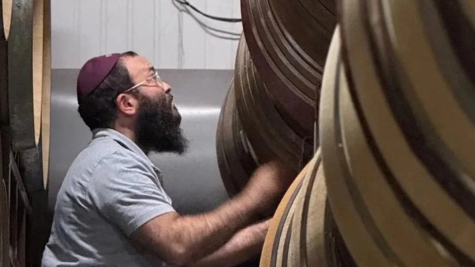 A man with glasses, a beard, and a purple kippah looks up and smiles while adjusting or inspecting large wooden or metal rings in a workshop or storage area.