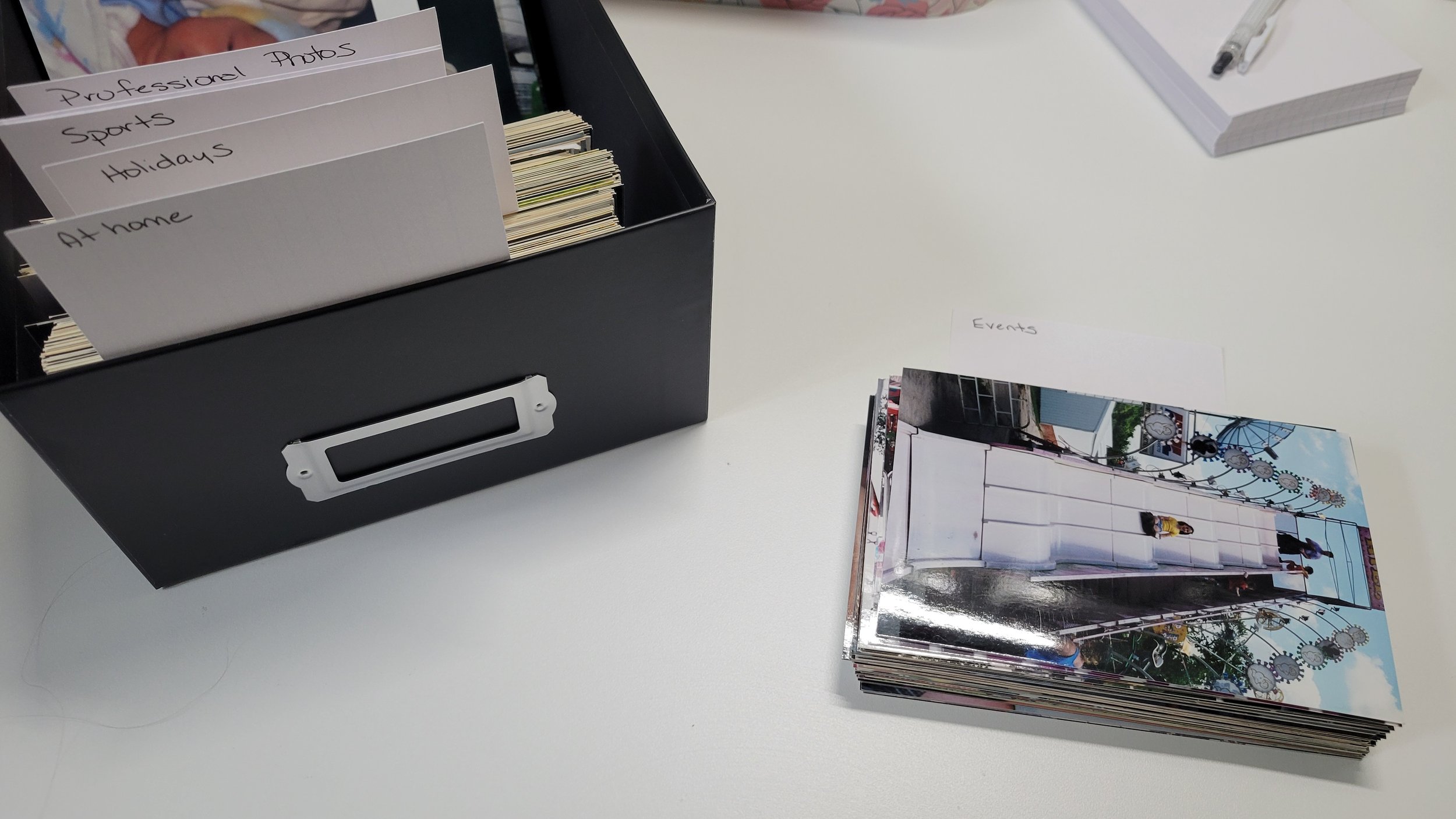 A black photo storage box with printed photos organized by handwritten category labels, with one photo pile set aside for events.