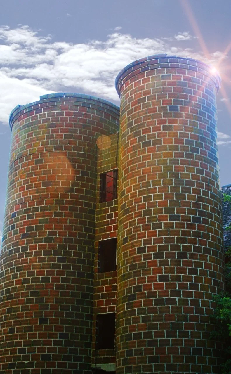 Two tall round brick towers photographed against a bright sky with sun flare.