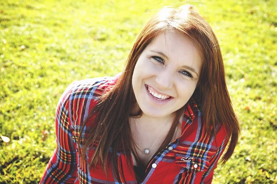 Senior girl smiling in casual outdoor portrait wearing red plaid shirt sitting on grass
