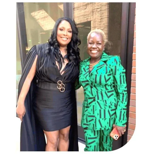 Two women smiling and standing together outside a building, one in a black dress with a cape and the other in a turquoise patterned suit.