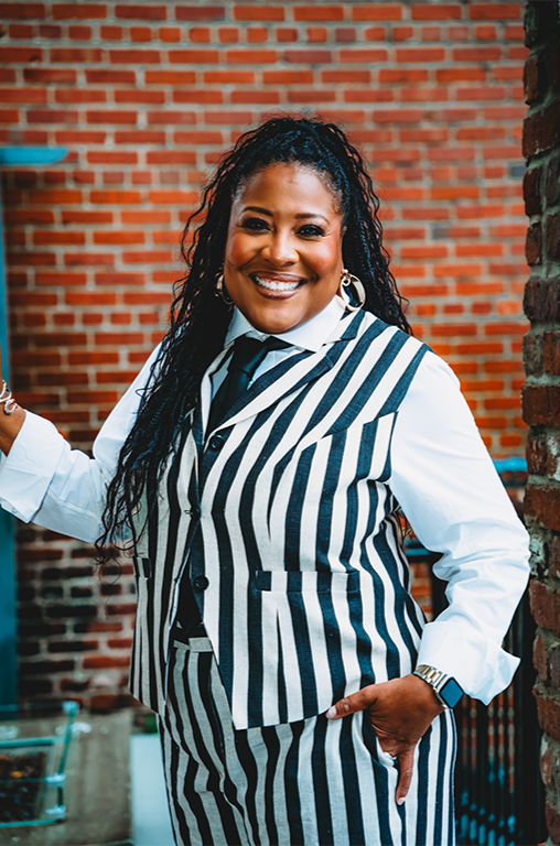 Smiling woman wearing a black and white striped vest and tie, white shirt, standing outdoors near a brick wall.