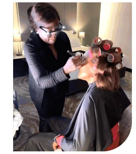 A makeup artist applying makeup on a woman sitting in a salon chair, with hair rollers in her hair, in a room with a sofa and lamps.