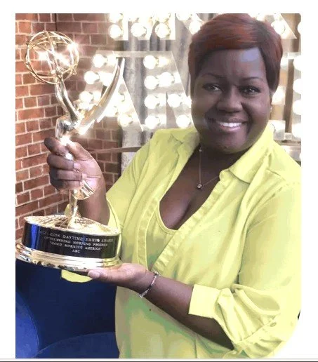 A woman with short hair smiling and holding a trophy in a well-lit room with a brick wall background.