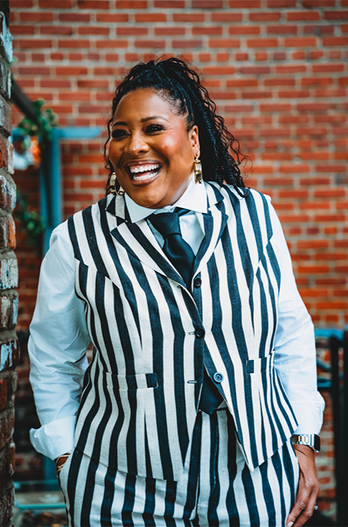 A woman in a striped vest and pants, with a white shirt and tie, smiling and standing outdoors against a brick wall.