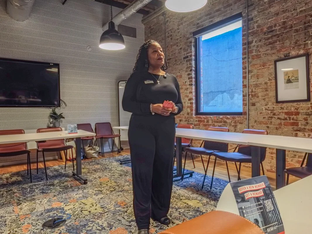 A woman stands in a conference room, holding a red object, surrounded by chairs and tables, with brick walls, a window, and a mounted TV.