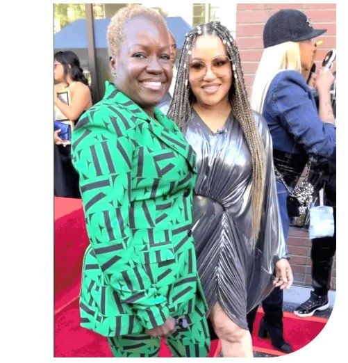 Two women standing and smiling at an outdoor event, one wearing a green patterned dress and the other in a shiny silver dress, with other people in the background.