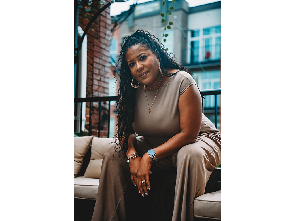 Woman with long curly hair wearing a beige outfit, sitting outdoors on a balcony with buildings in the background.