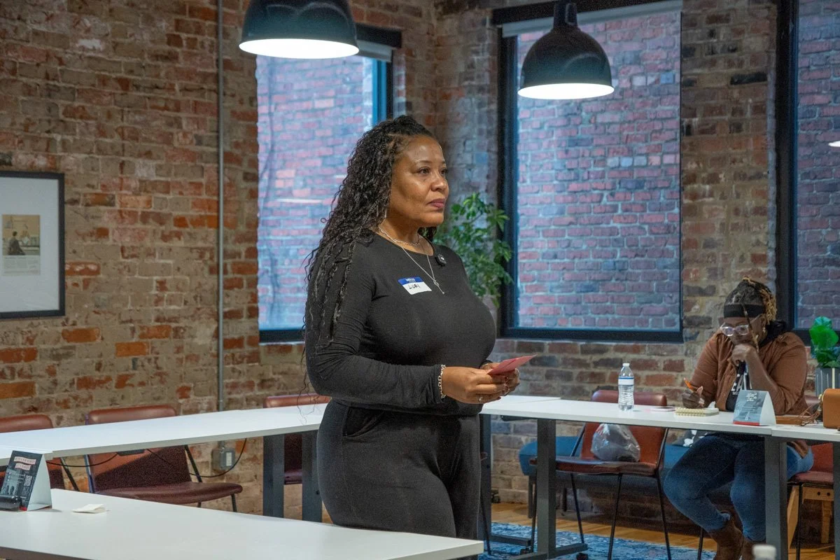 A woman standing in a meeting room, holding a red piece of paper, wearing a black outfit, with a name tag, in front of a brick wall and windows, with another person sitting at the table.