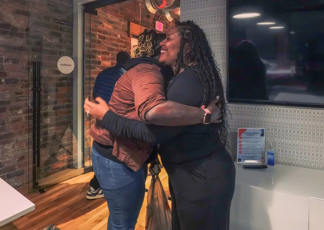 Two women hugging inside a room with brick walls and a television mounted on the wall.