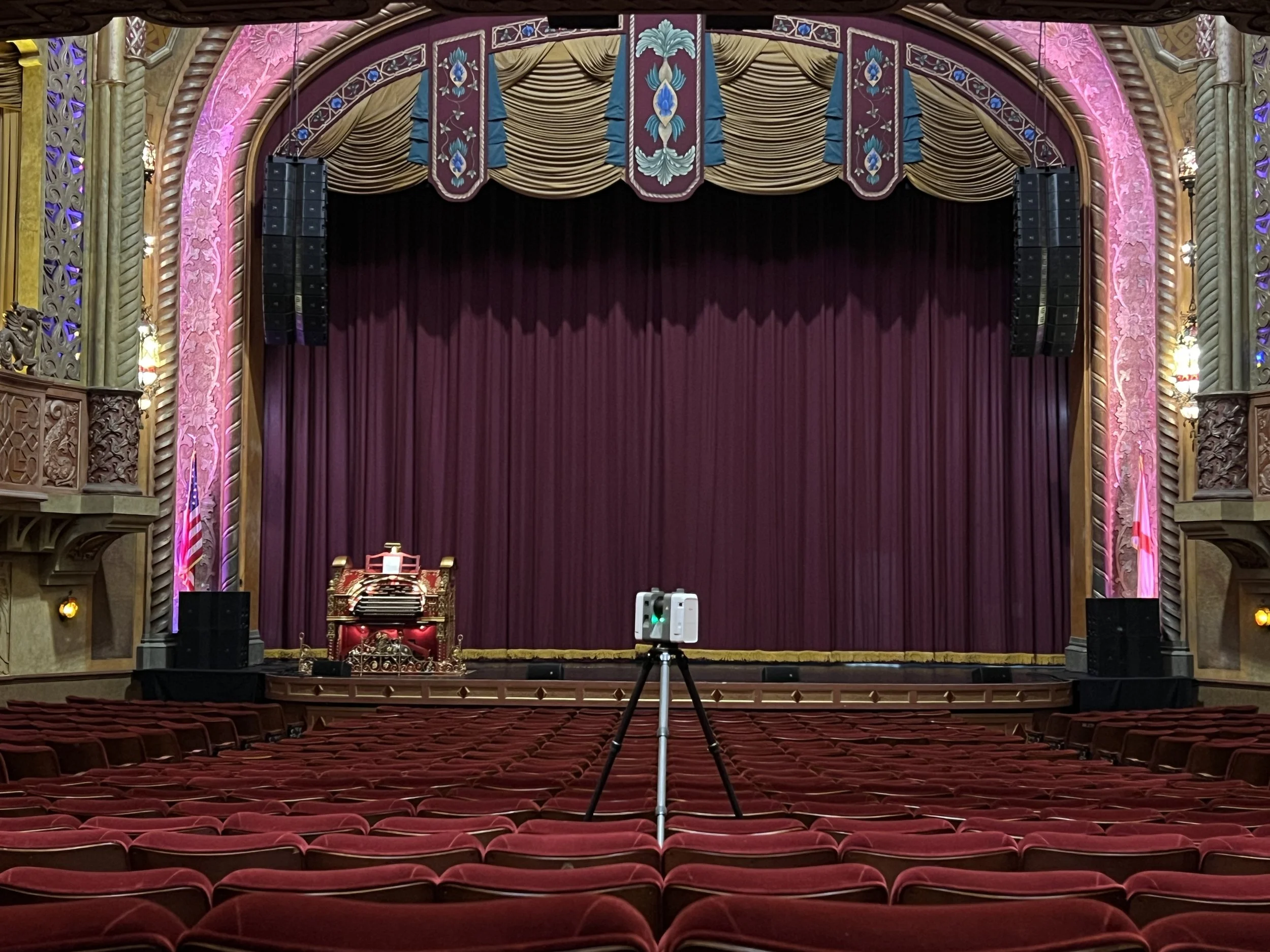 An empty theater stage with maroon curtains and elaborate gold, pink, and purple decor, stage lighting, and a large speaker system on either side. Rows of red velvet seats extend toward the stage.