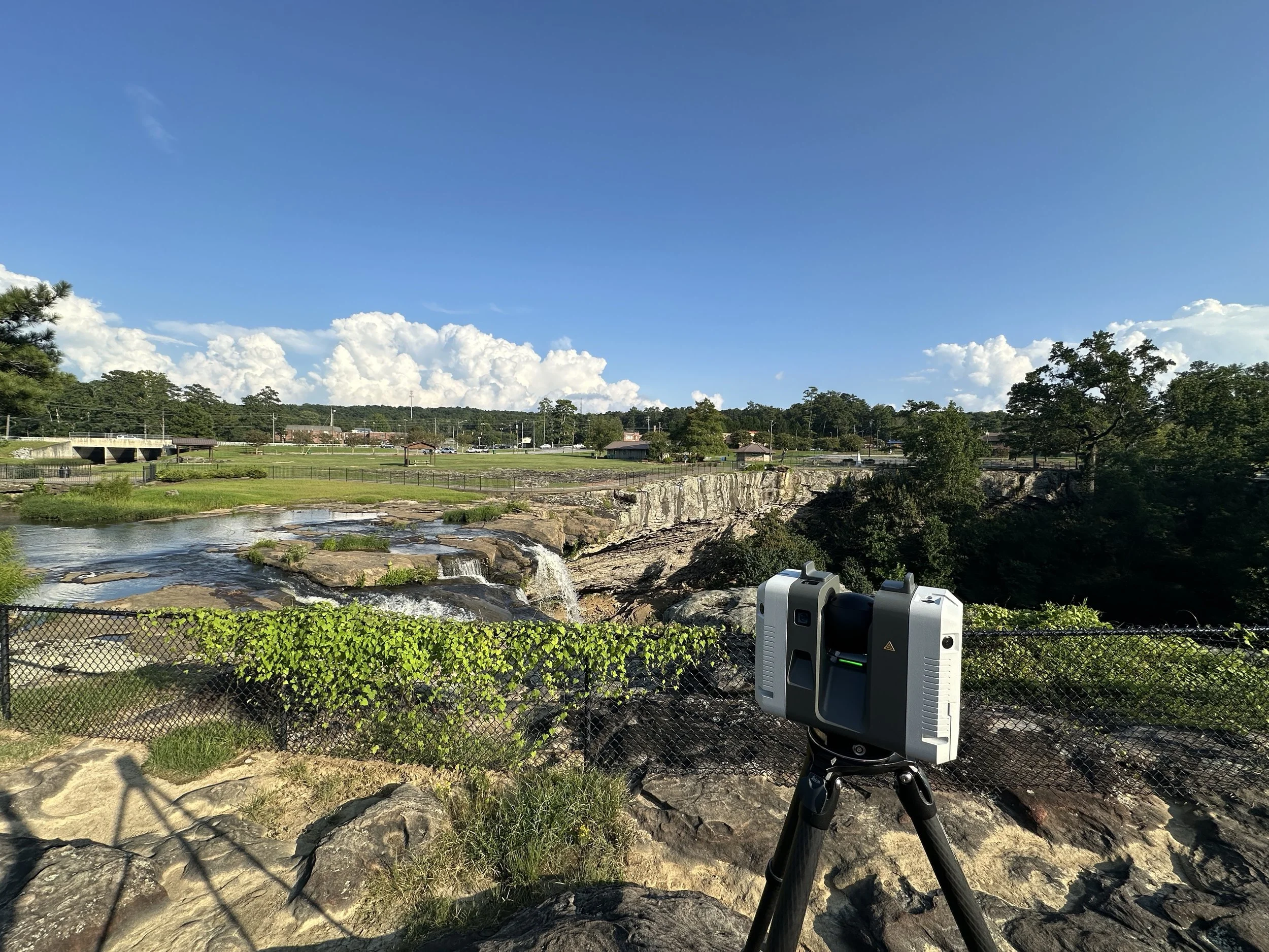 A scenic view of a park with a river, rocks, trees, and a blue sky with clouds. The RTC360 scanner is set up in the foreground.