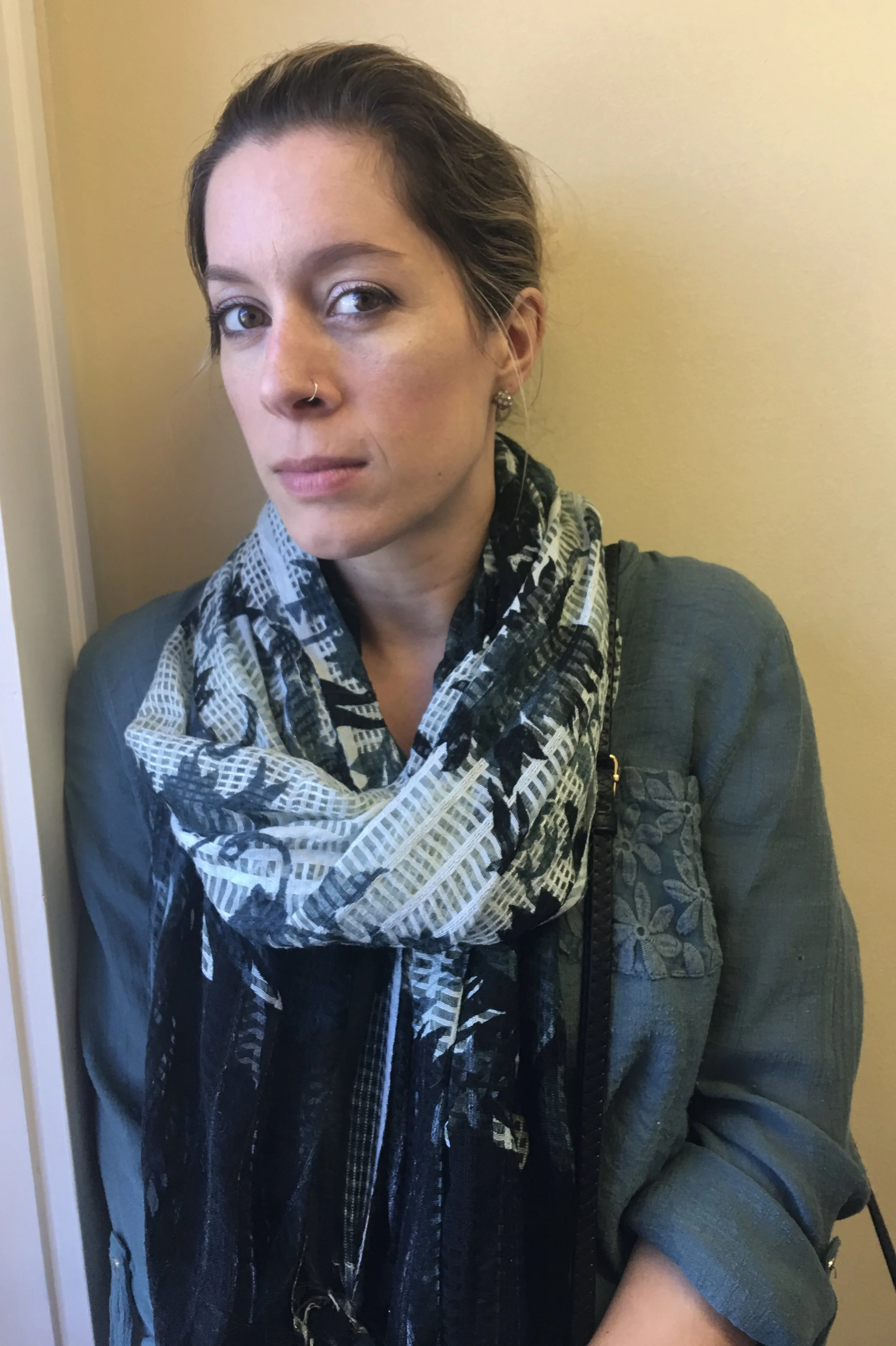 A woman with light skin, short brown hair, wearing a dark green jacket, patterned scarf, and earrings, standing indoors against a beige wall.
