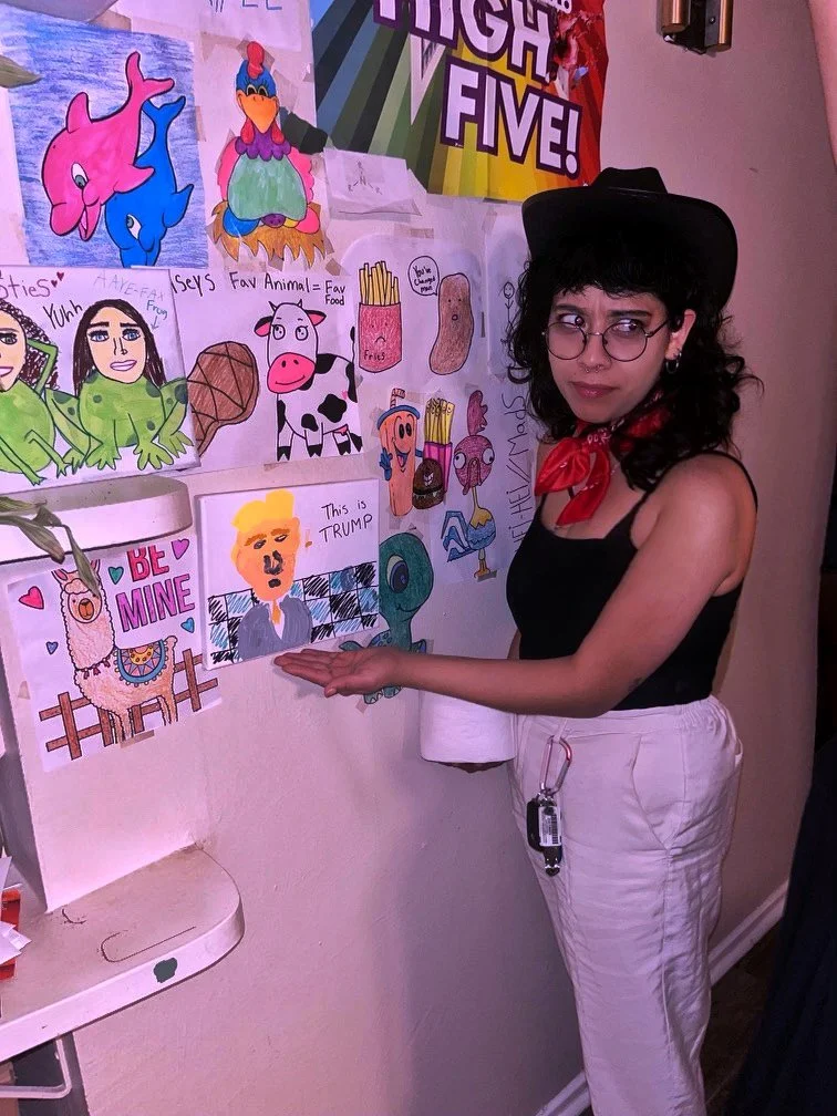 A woman with dark hair, glasses, and earrings, dressed in a black top and white pants, stands in front of a colorful wall of drawings and posters, pointing to a drawing of Donald Trump with a sign that says "This is TRUMP".