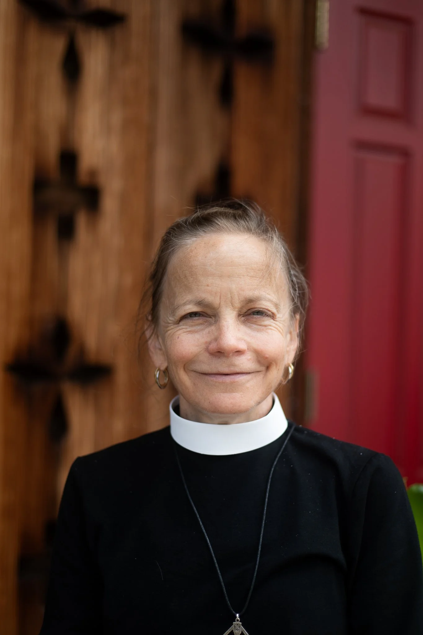 A middle-aged woman wearing a black clergy shirt and a white clerical collar, smiling outdoors with a wooden wall and red door in the background.