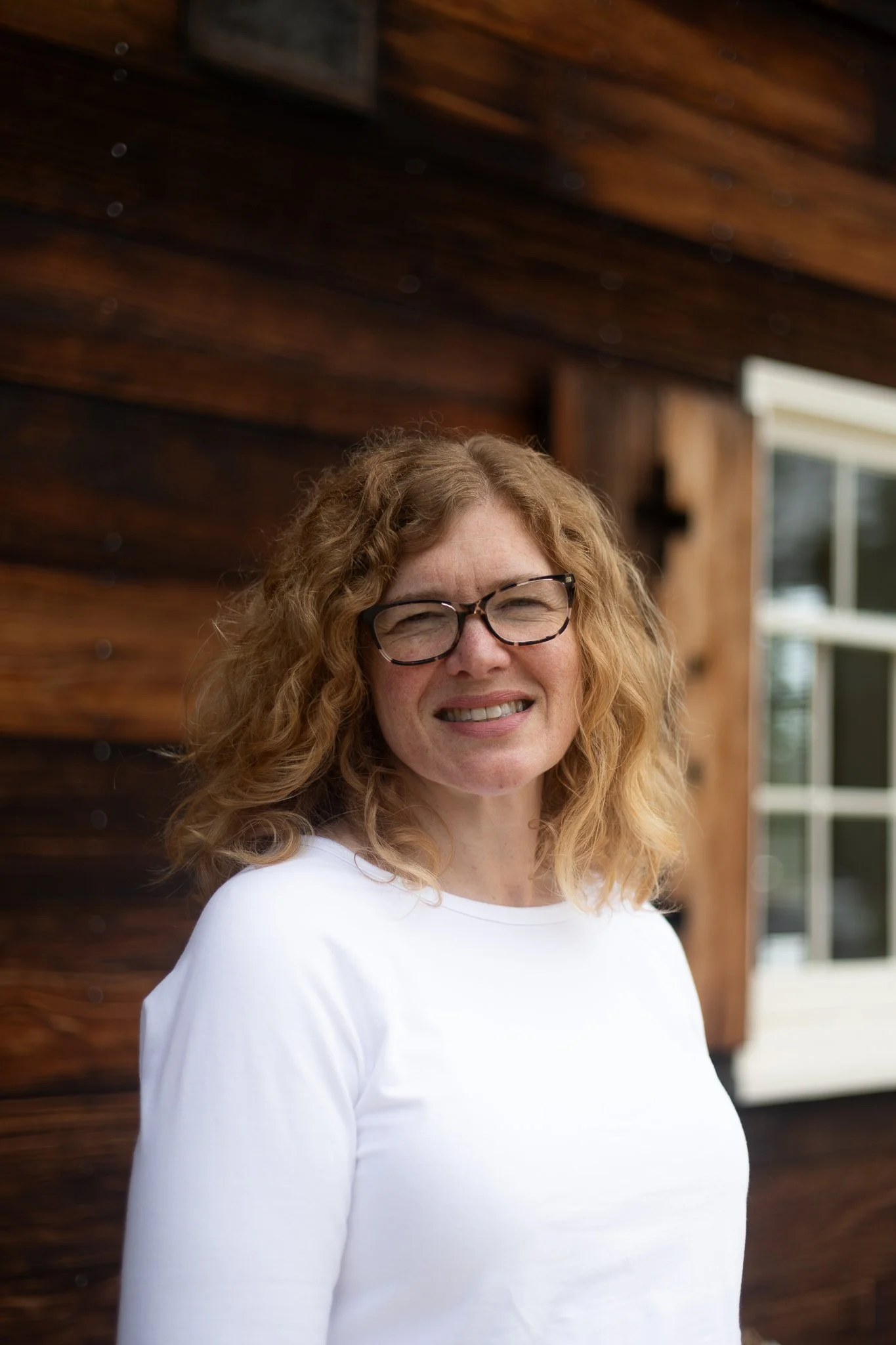 A woman with curly red hair and glasses is smiling, standing outside near a rustic wooden building with a window.