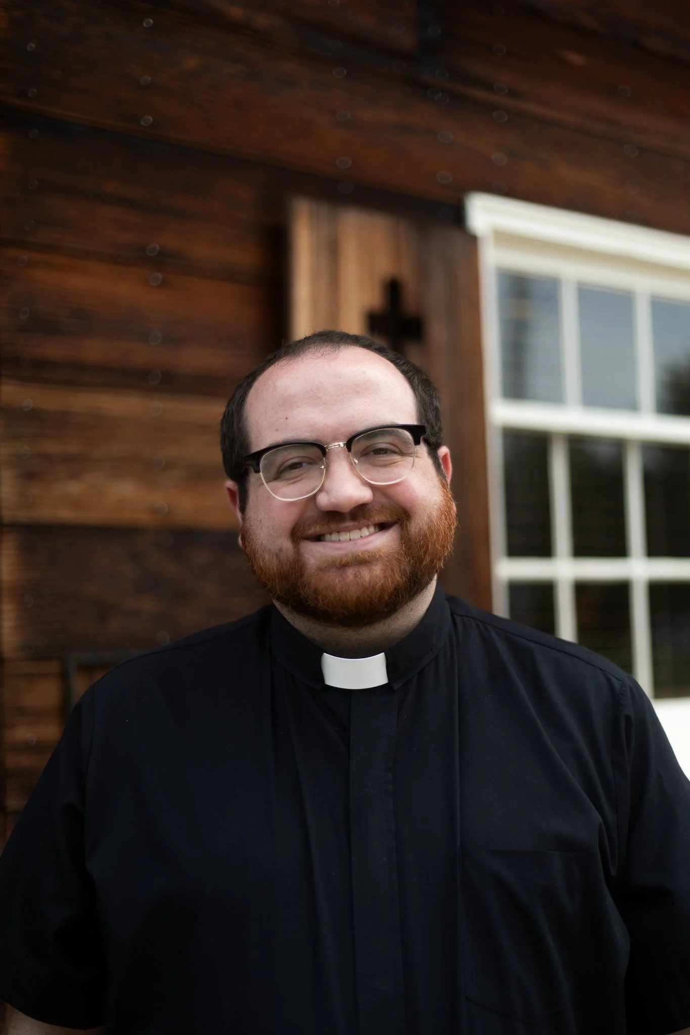 A smiling man dressed as a priest standing in front of a wooden house with a cross on the wall and a window.