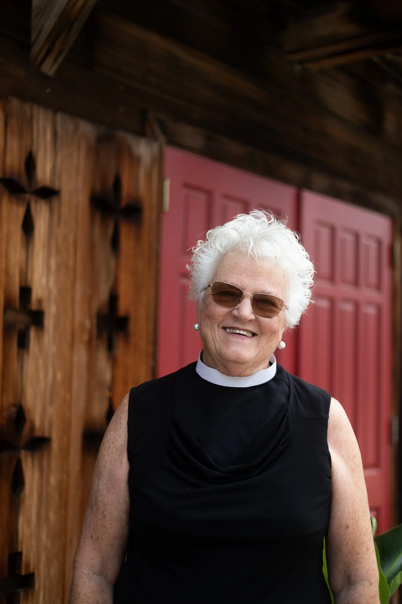 An elderly woman with white curly hair, wearing sunglasses, pearl earrings, and a black sleeveless top with a white clerical collar, smiling outdoors in front of a wooden wall and red door.