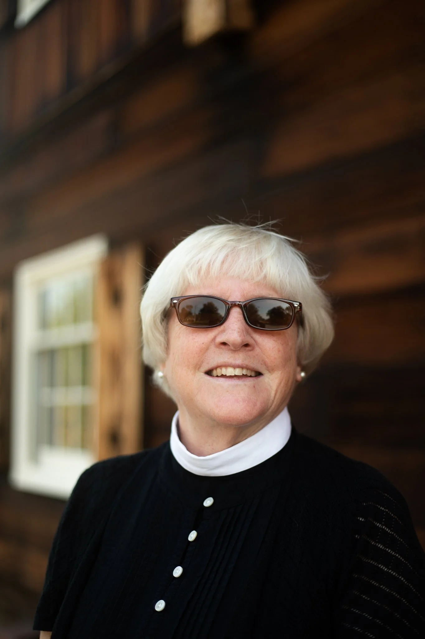 An elderly woman with short white hair, wearing sunglasses and a black top with a white collar, smiling outdoors in front of a wooden building.
