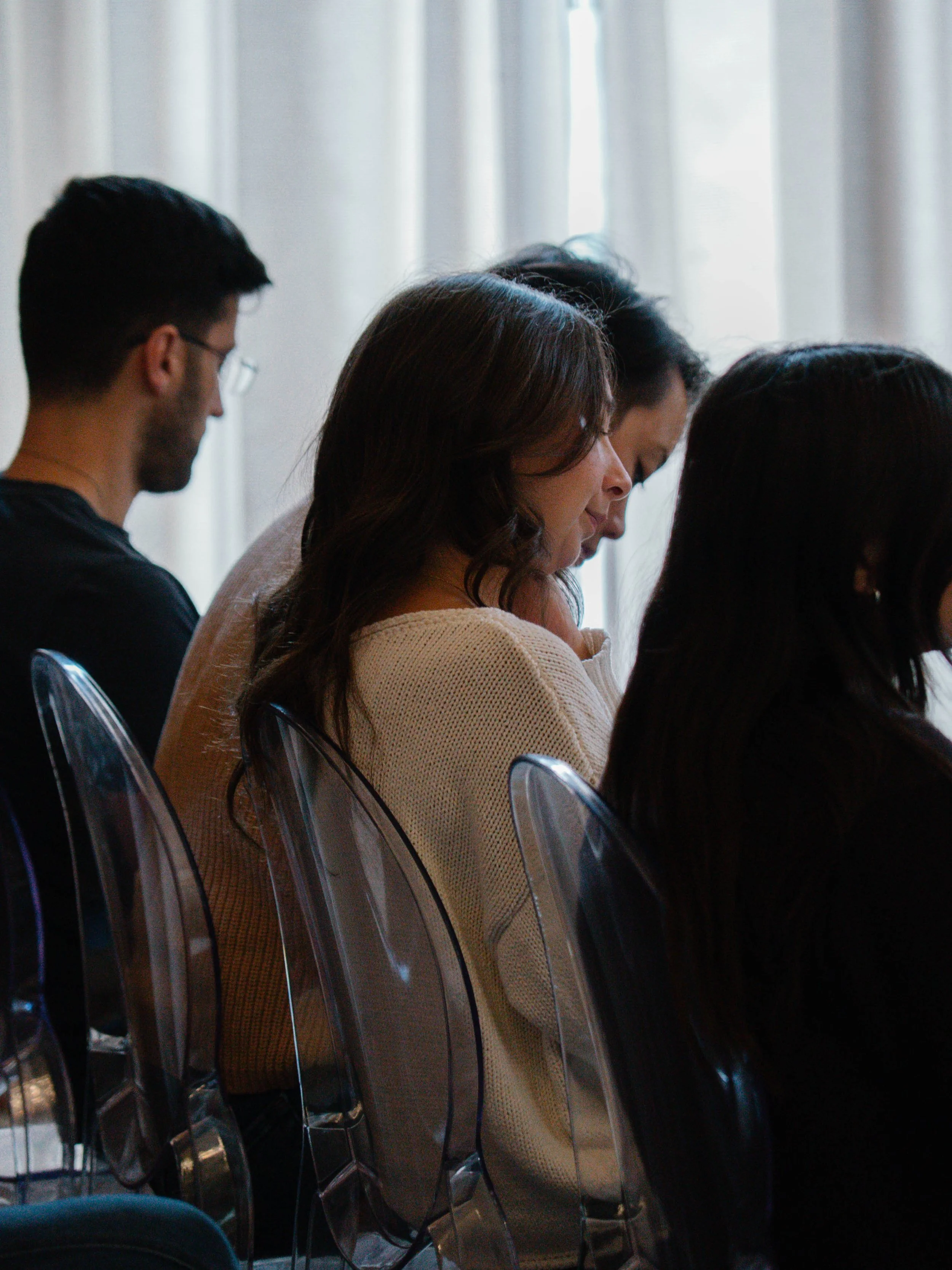 Four individuals seated next to each other, appearing to be engaged or concentrating, with light coming through window blinds in the background.
