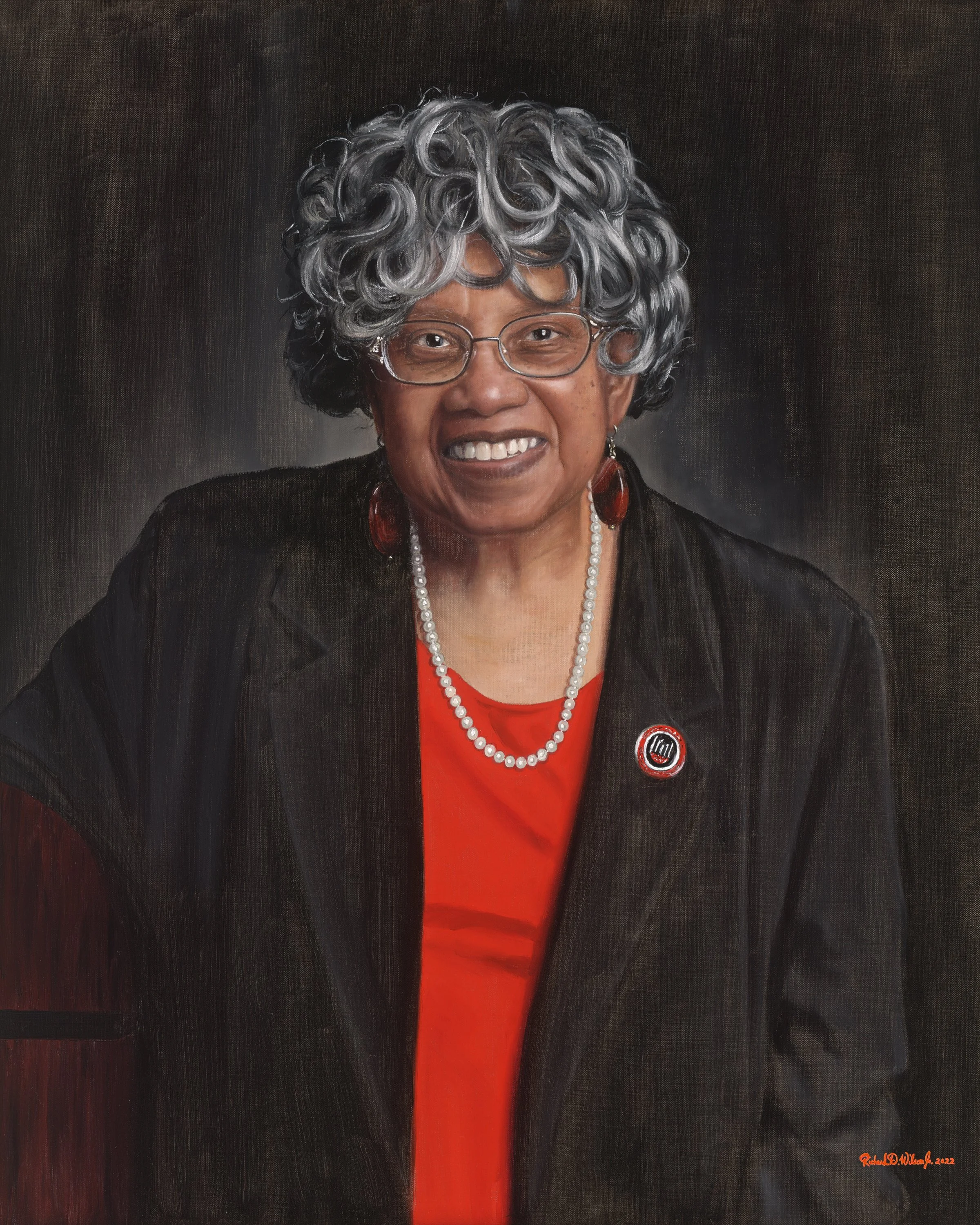 Portrait of an elderly African American woman with gray curly hair wearing glasses, a red top, a black jacket, pearl necklace, red earrings, and a pin, smiling against a dark background.