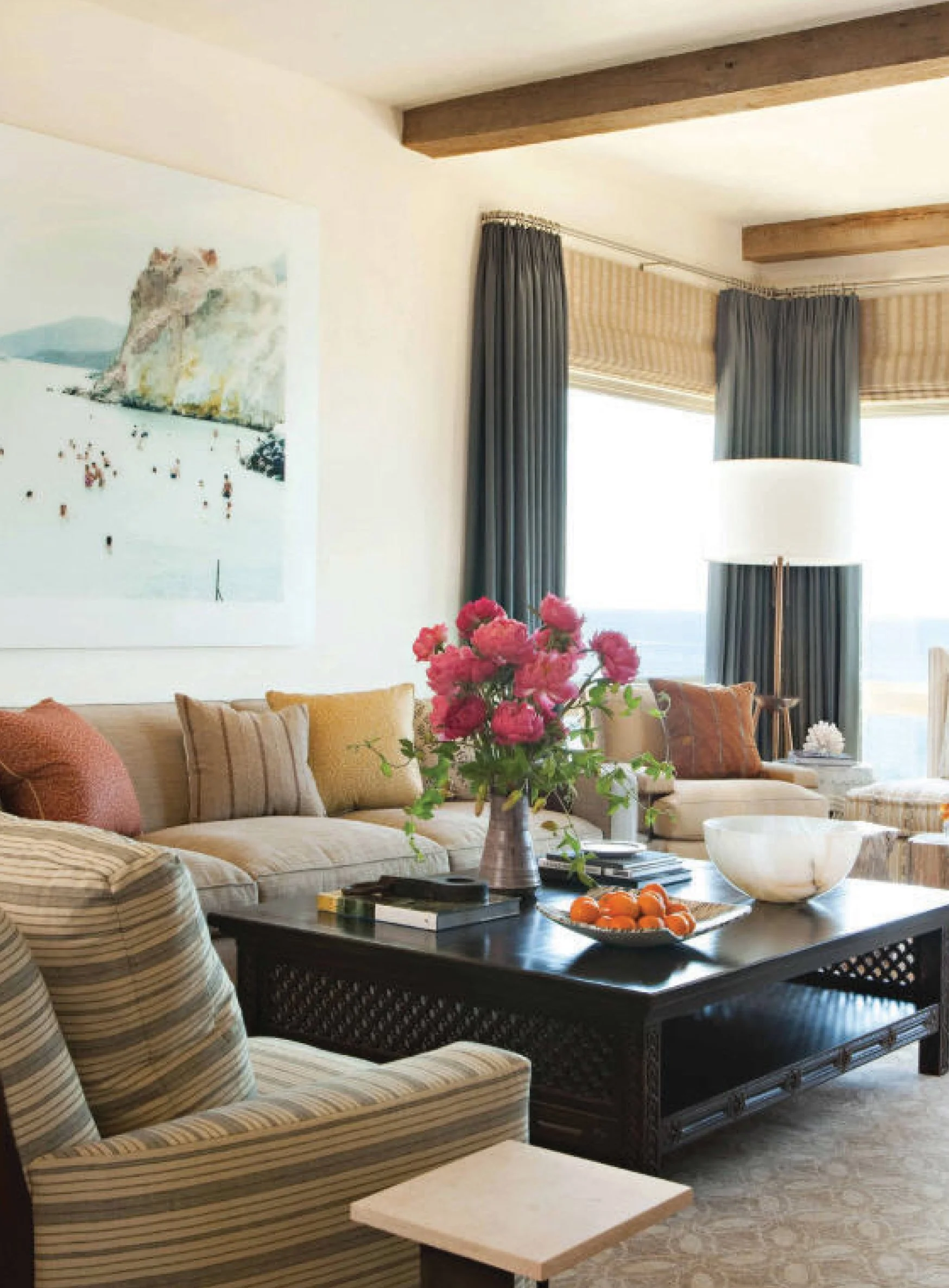 Living room with beige sofa, striped armchair, black coffee table holding a pink flower vase and a bowl of oranges, window with blue curtains, wall art, and floor lamp.