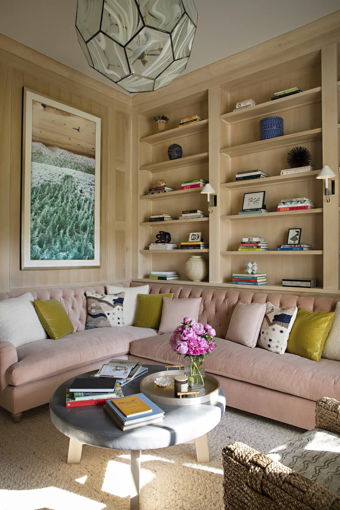 Cozy sitting room by Cameron Design featuring a blush tufted sectional with patterned and velvet accent pillows, built in light oak bookshelves styled with art and books, and a round gray coffee table topped with flowers and decor. A large aerial bea