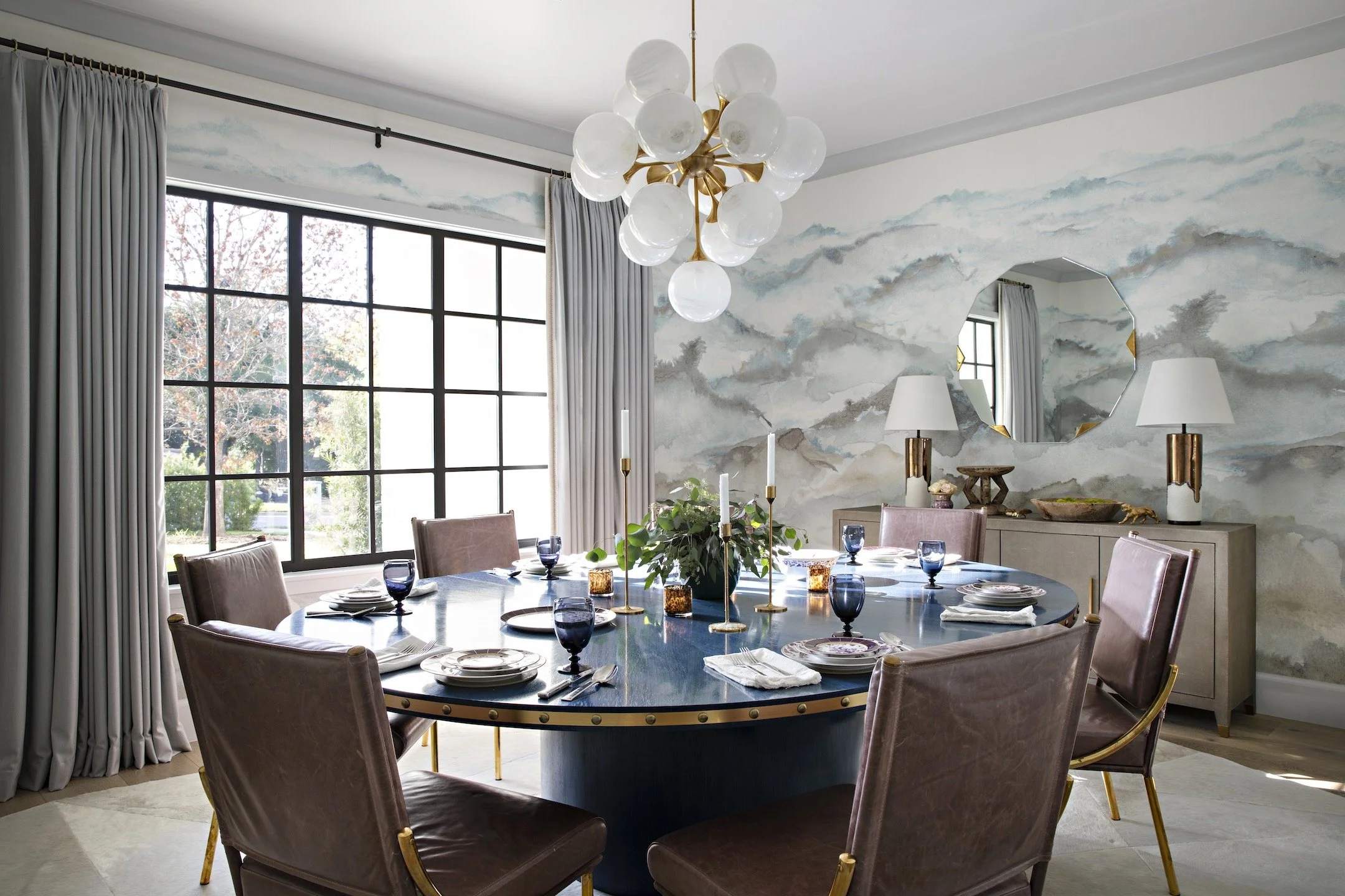 Elegant dining room with a large round table set for eight, featuring white plates, silverware, and blue glassware, surrounded by upholstered chairs with gold accents. A modern chandelier with multiple glass globes hangs above the table. The room has