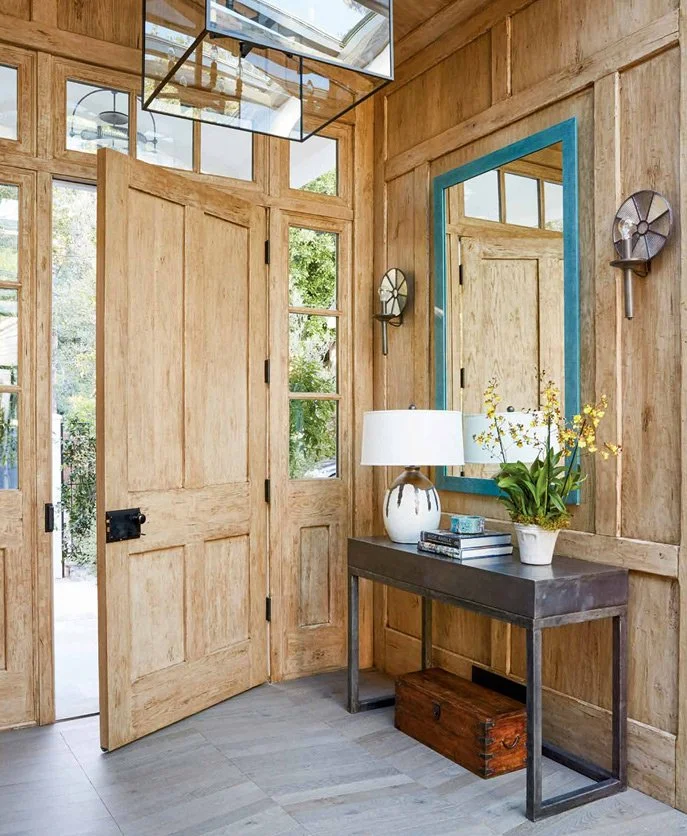 Cameron Design foyer with light wood paneled doors, a modern console table, blue-trimmed mirror, and a table lamp with fresh flowers.