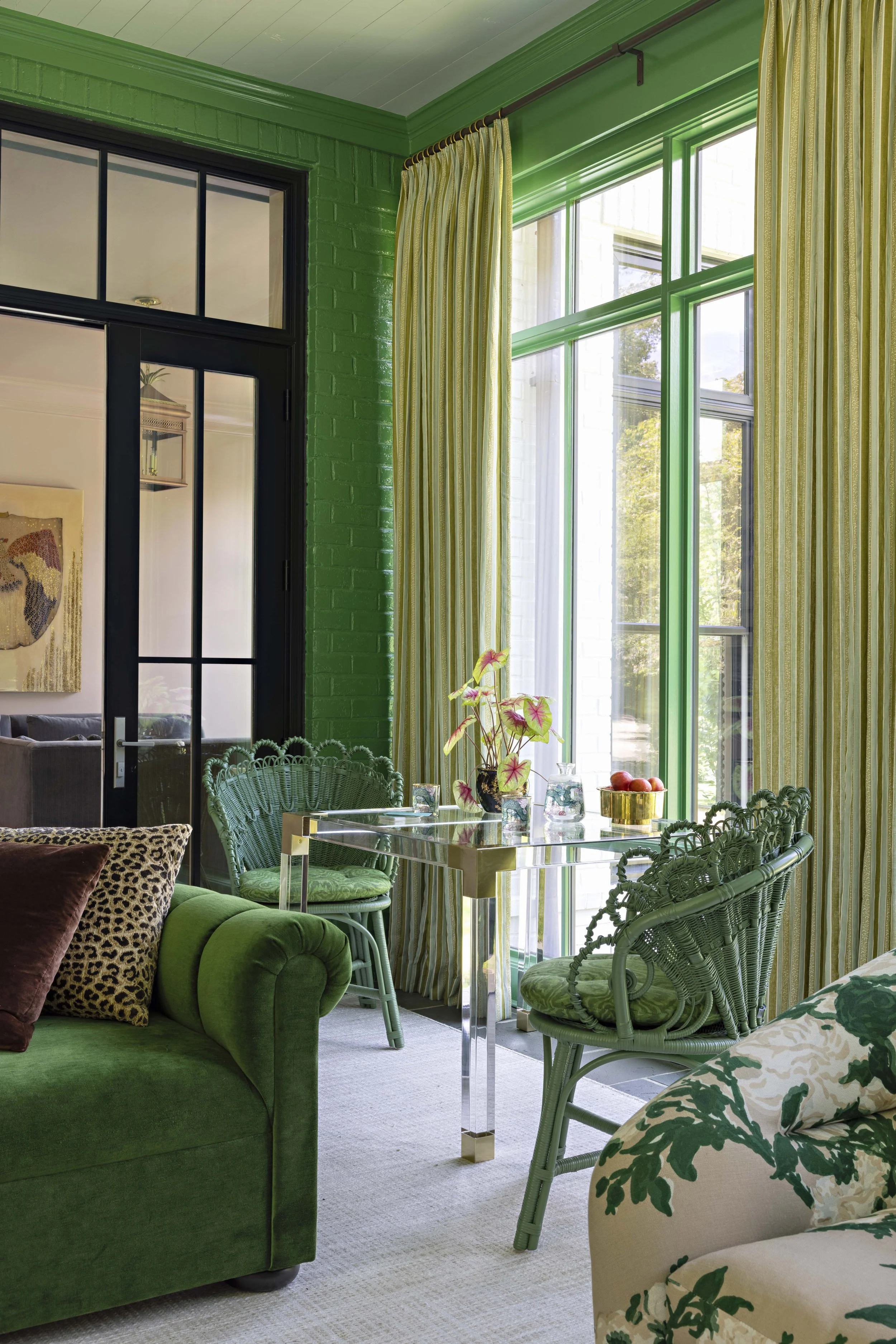 A cozy living room with green furniture, yellow-striped curtains, and large windows allowing natural light, decorated with plants on a glass table.