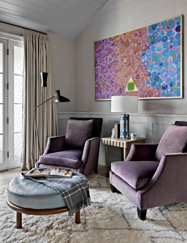 A cozy reading corner with two deep purple velvet armchairs, a round ottoman with a blue cushion and plaid throw, a textured rug, and a side table with books and a blue ceramic lamp. Large modern artwork with geometric patterns in pink, purple, and b