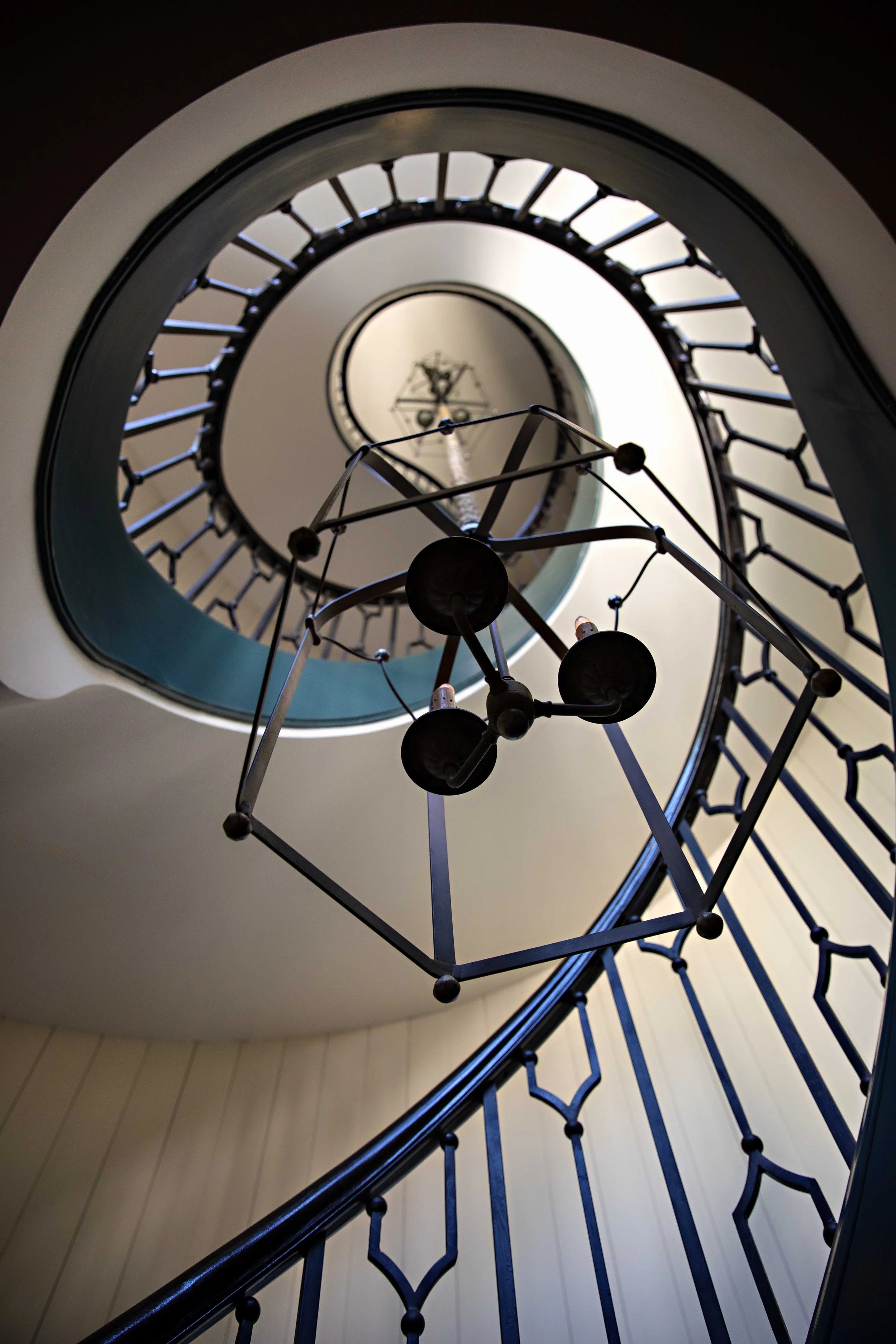 Looking up a spiral staircase with decorative white walls and a black metal handrail, featuring a modern geometric chandelier hanging in the center.