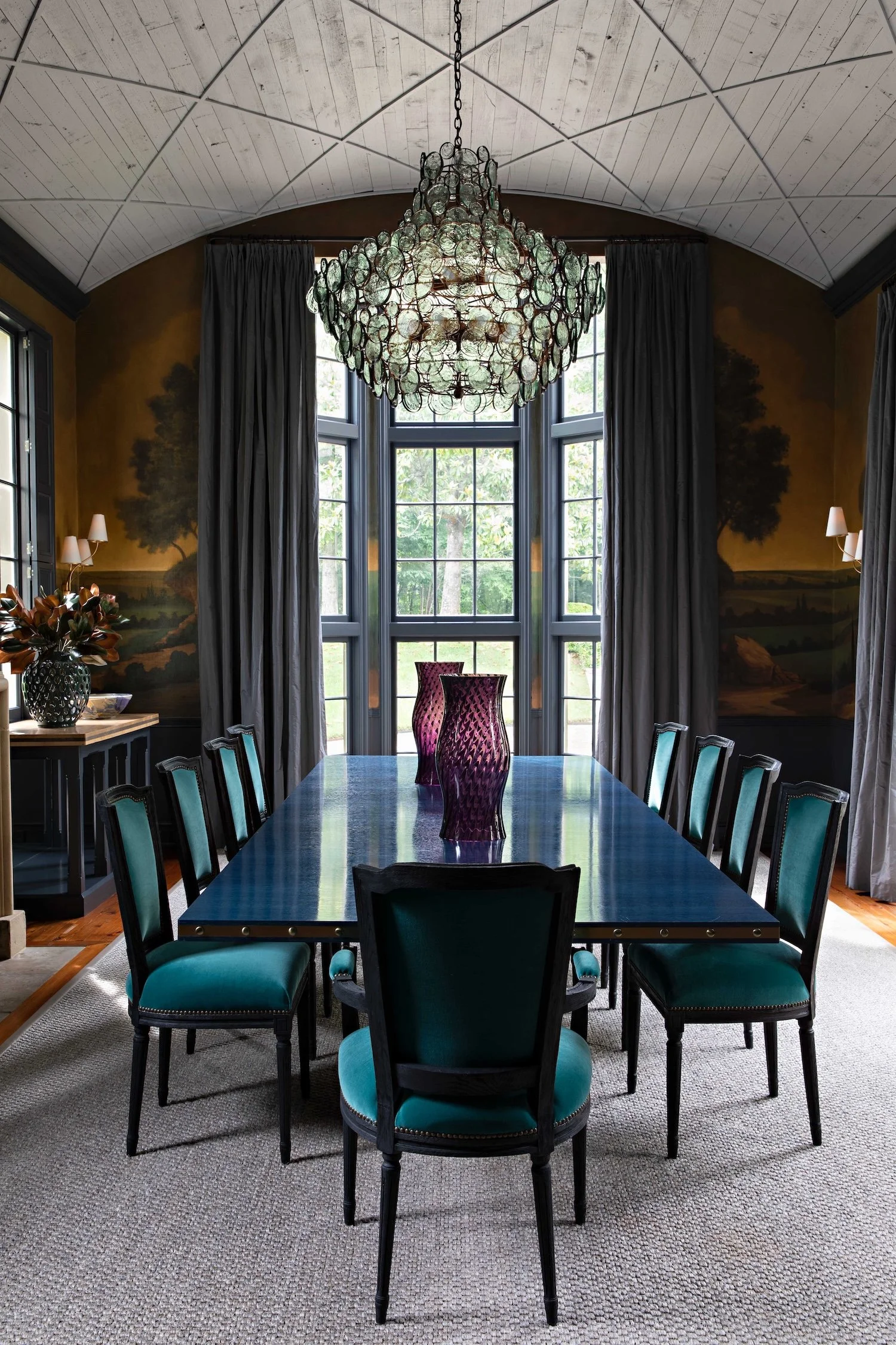 Elegant dining room with a large, polished table surrounded by ten teal-upholstered chairs, a decorative chandelier hanging from a vaulted ceiling, tall windows with gray curtains, and wall paintings of trees.